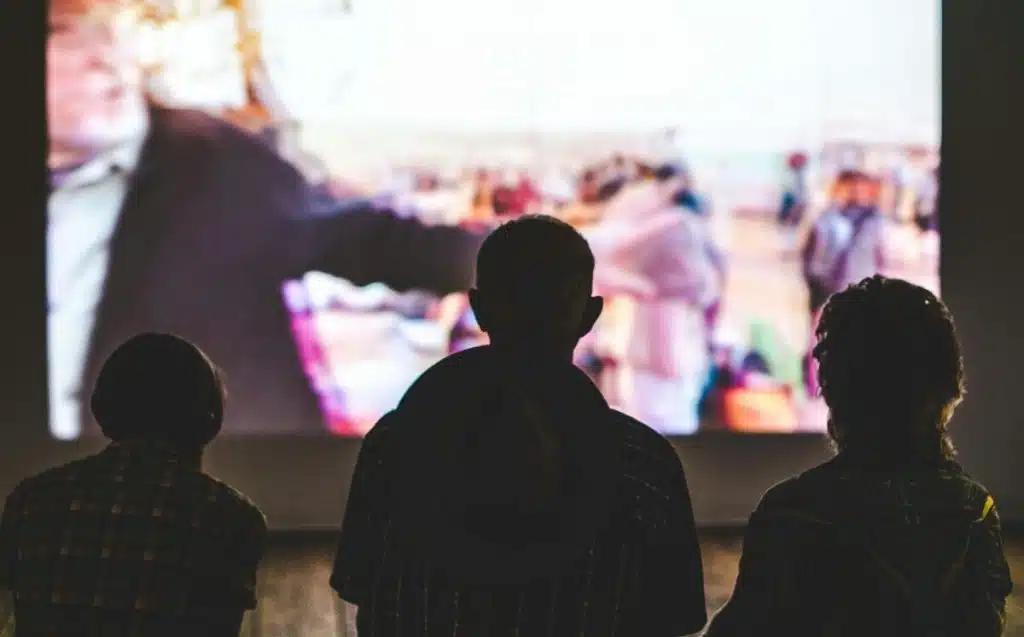 Im Hintergrund läuft ein Film auf einer Leinwand, vorne sind schemenhaft Personen zu sehen.