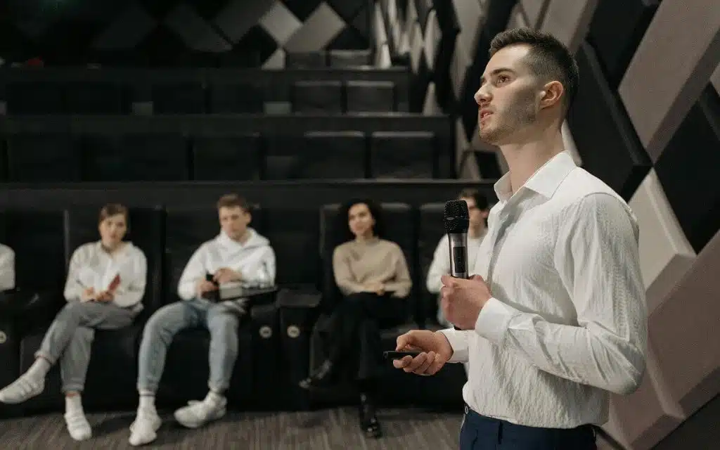 Ein Schauspieler mit einem Mikrofon steht vor einer Gruppe sitzender Personen