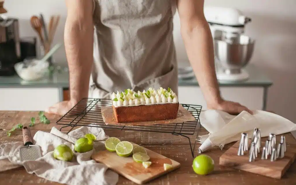 Fertig gebackener Kastenkuchen auf Kuchengitter, dekoriert mit Limettenscheiben. Im Hintergrund eine Person mit Schürze