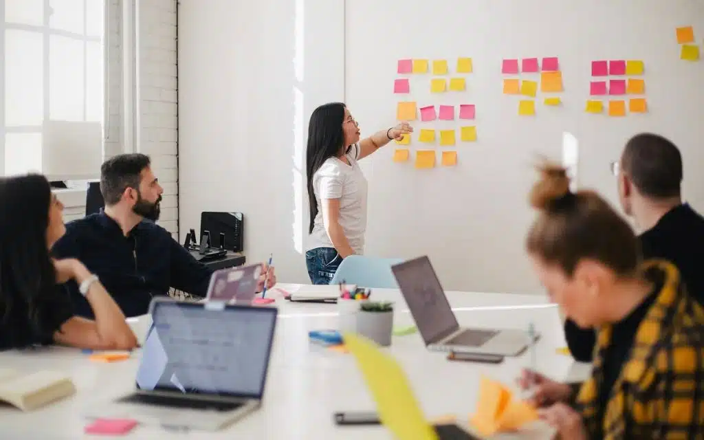 Frau, die einen Vortrag hält steht vor einer Gruppe Menschen, die um einen Tisch sitzt. Im Hintergrund hängen an der Wand bunte Post-its
